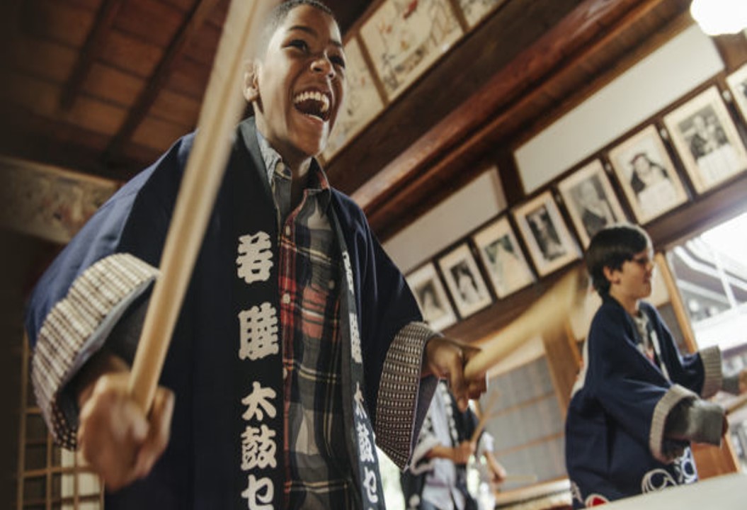 3 boys on a family trip to Japan, dressed in traditional robes and happily playing ceremonial drums.