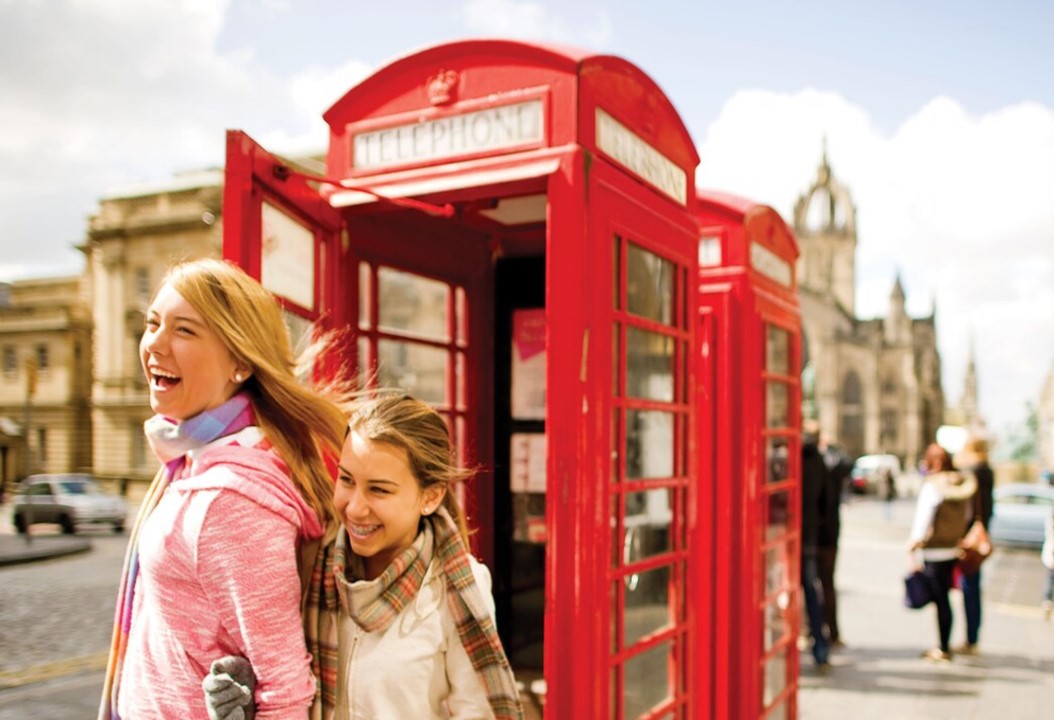 Two light-brown haired teenage sisters exiting a London's iconic red telephone booths with wide smiles on their face.