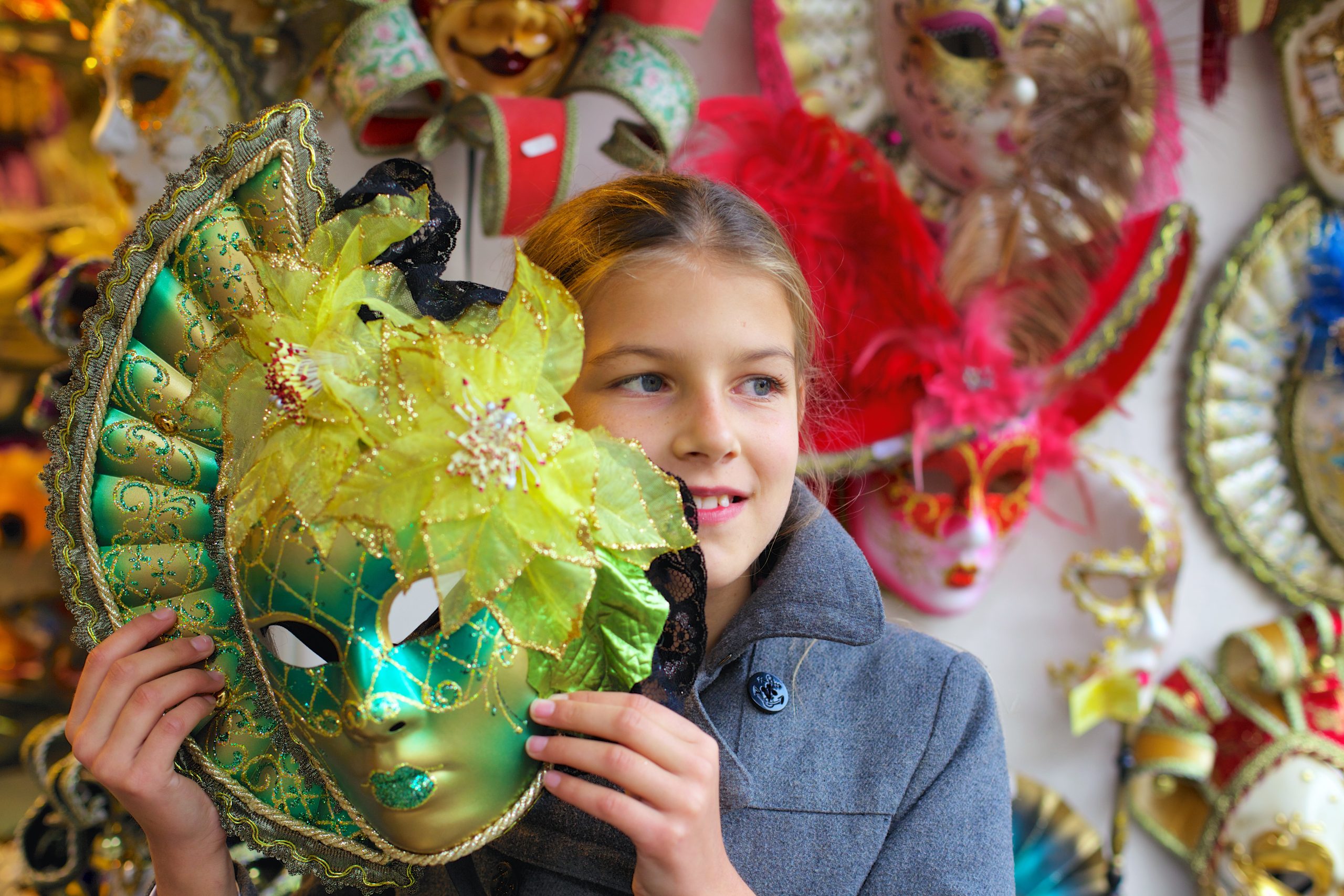 blonde pre-teenaged girl holds a lime green masquerade ball mask. In the background is a wall full of similar masks in pink, gold, and red.