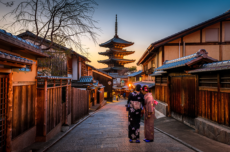 two women dressed in traditional geisha wear standing in the middle of a quiet old town street in Kyoto