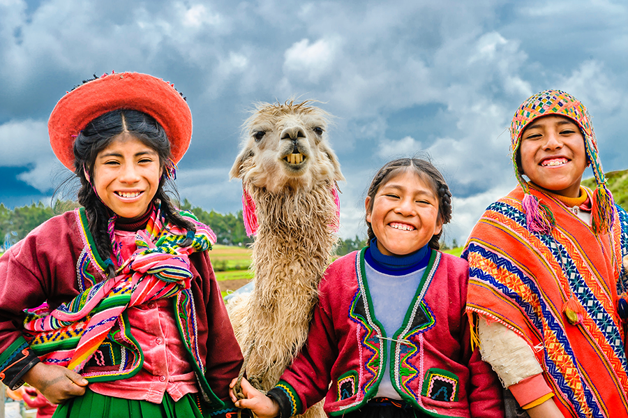 three smiling pervuian children dressed in traditional wear as they pose with a half-smiling llama